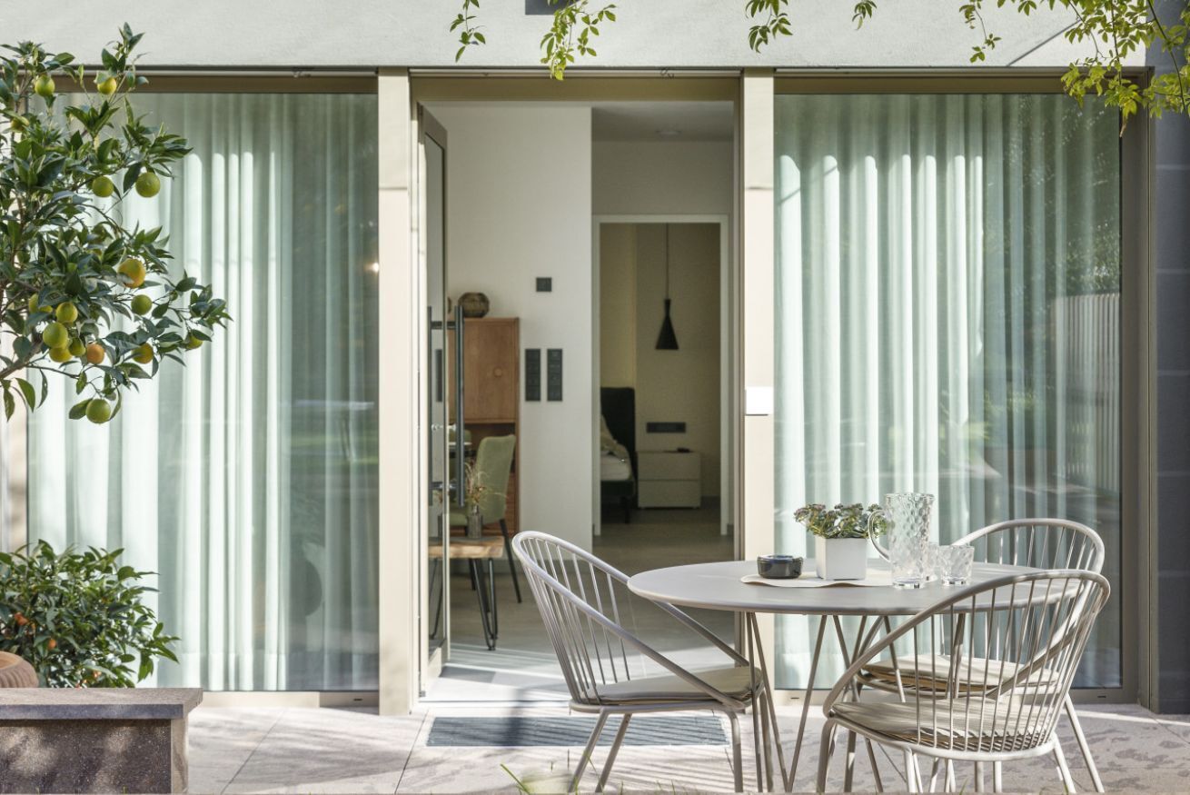Un espace repas extérieur moderne, avec une table et des chaises, une plante en pot et une vue sur l’intérieur de la maison à travers une porte vitrée.