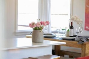 Un comedor con una mesa dispuesta para dos personas, un jarrón con flores rosadas y una botella de vino sobre la mesa.