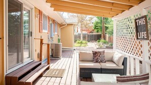 Un patio couvert avec un banc en bois, un canapé blanc avec des coussins et un mur en treillis orné d’une enseigne.