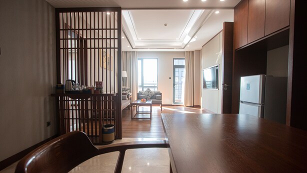 A modern kitchen with wooden cabinets, a dining area with a table and chairs, and a view of the living room through a sliding door. - VIENTIANMEKONGHOTEL (Vientiane)