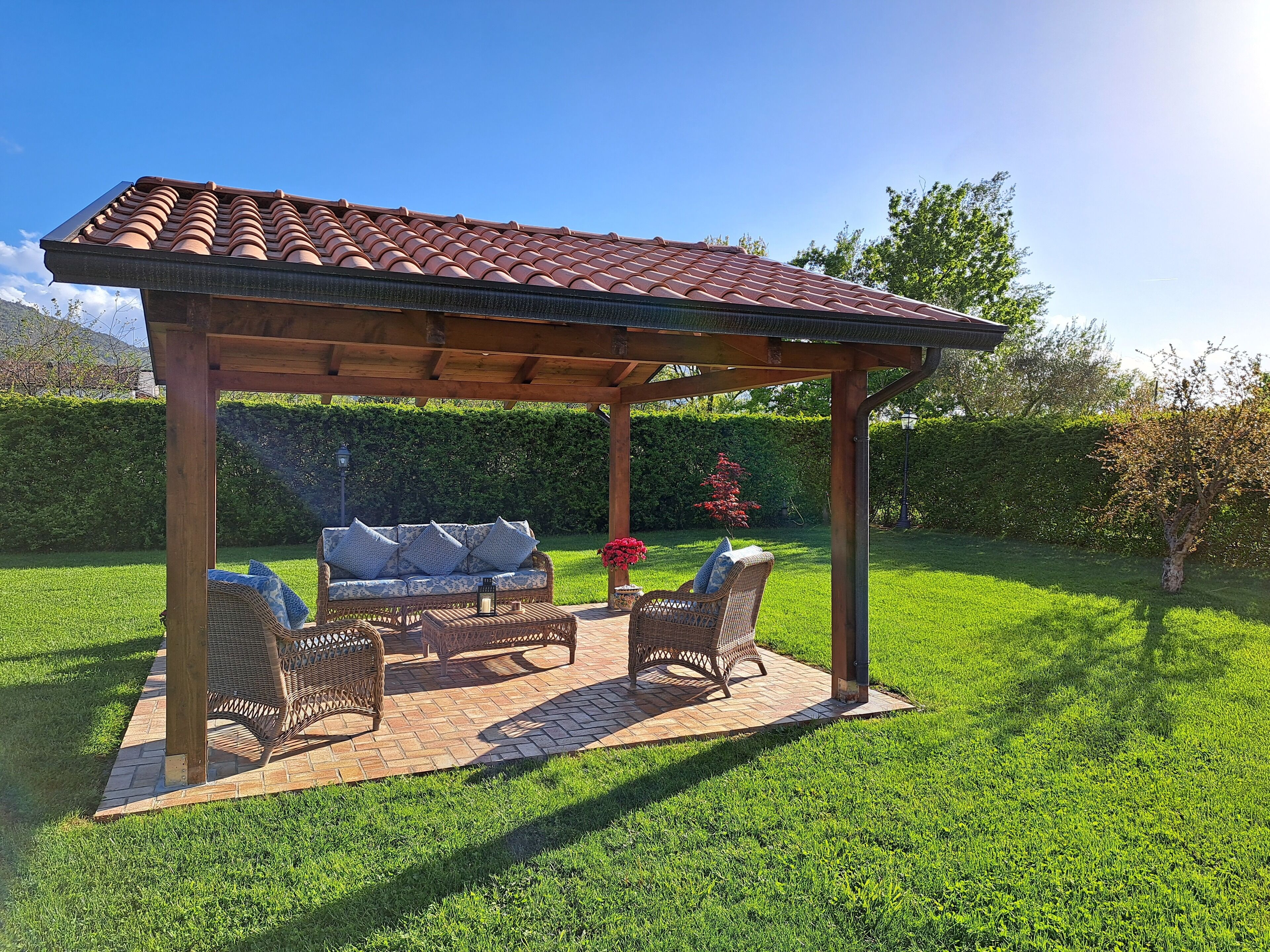 Une pergola en bois surmontée d’un toit en tuiles, un canapé en osier et deux fauteuils en osier, le tout dans un jardin bien entretenu.