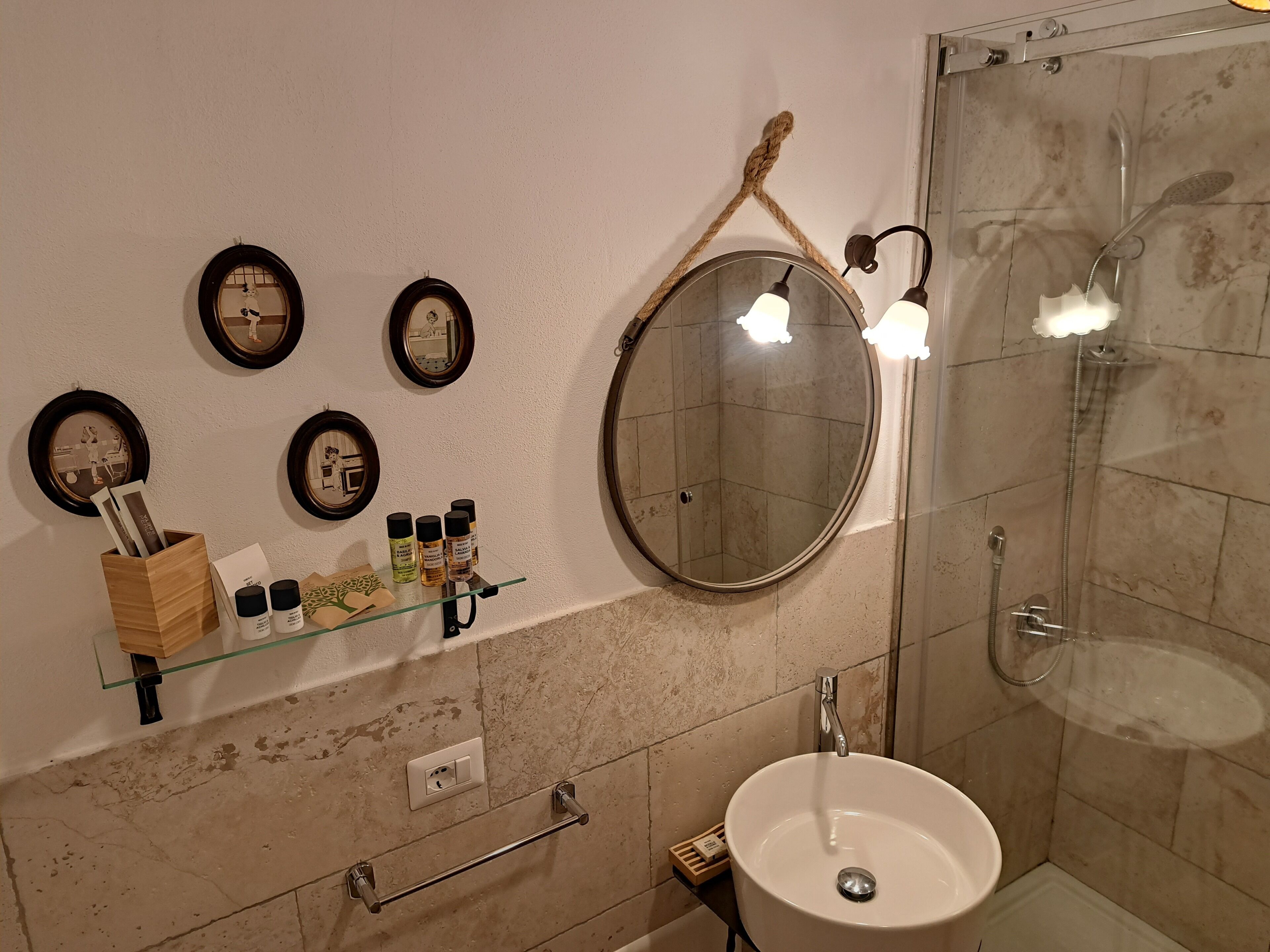 A bathroom with a round mirror, a glass shelf with toiletries, a sink, and a shower.