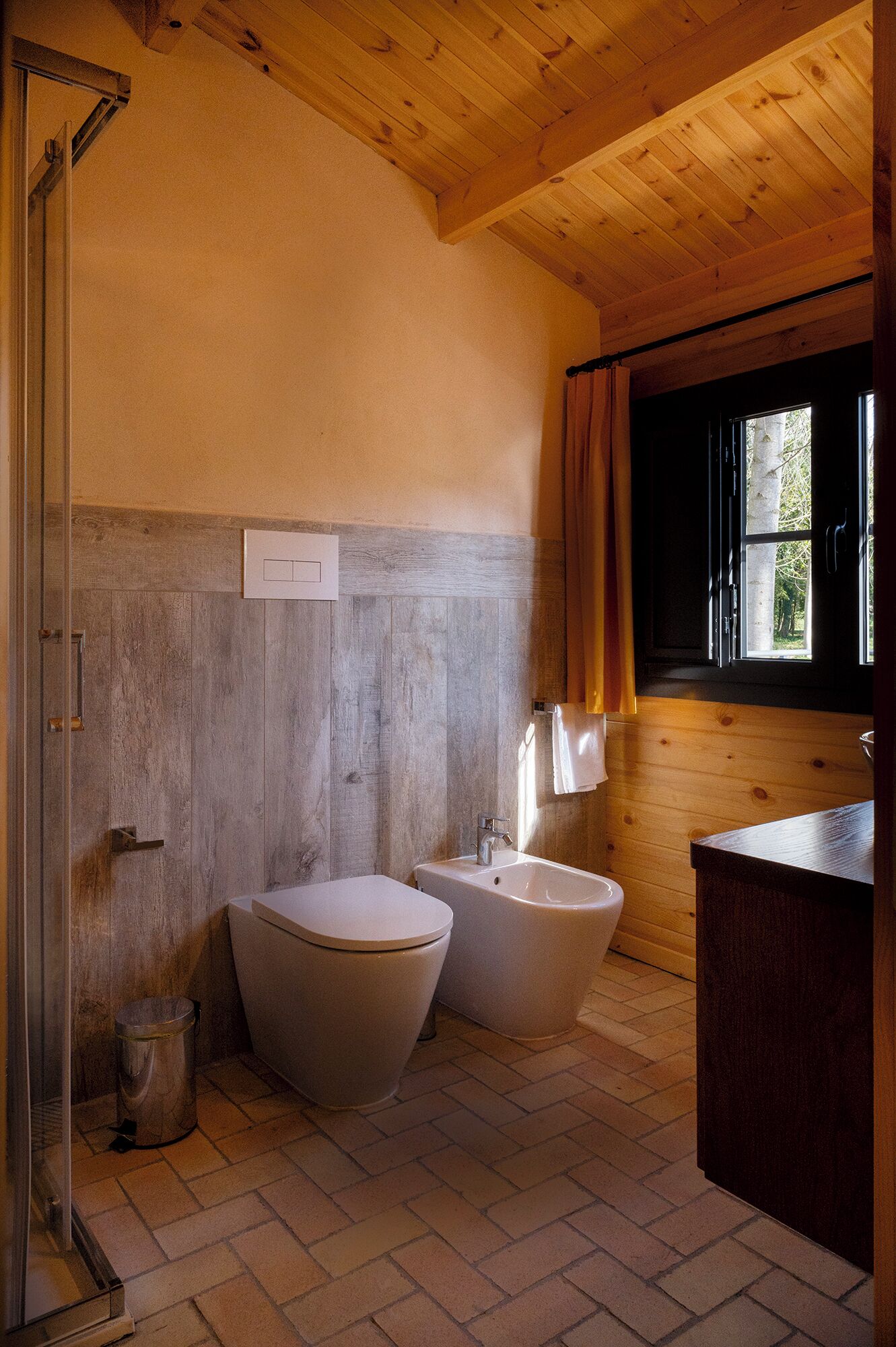 A bathroom with a wooden ceiling, two toilets, a bidet, a sink, and a window with curtains.