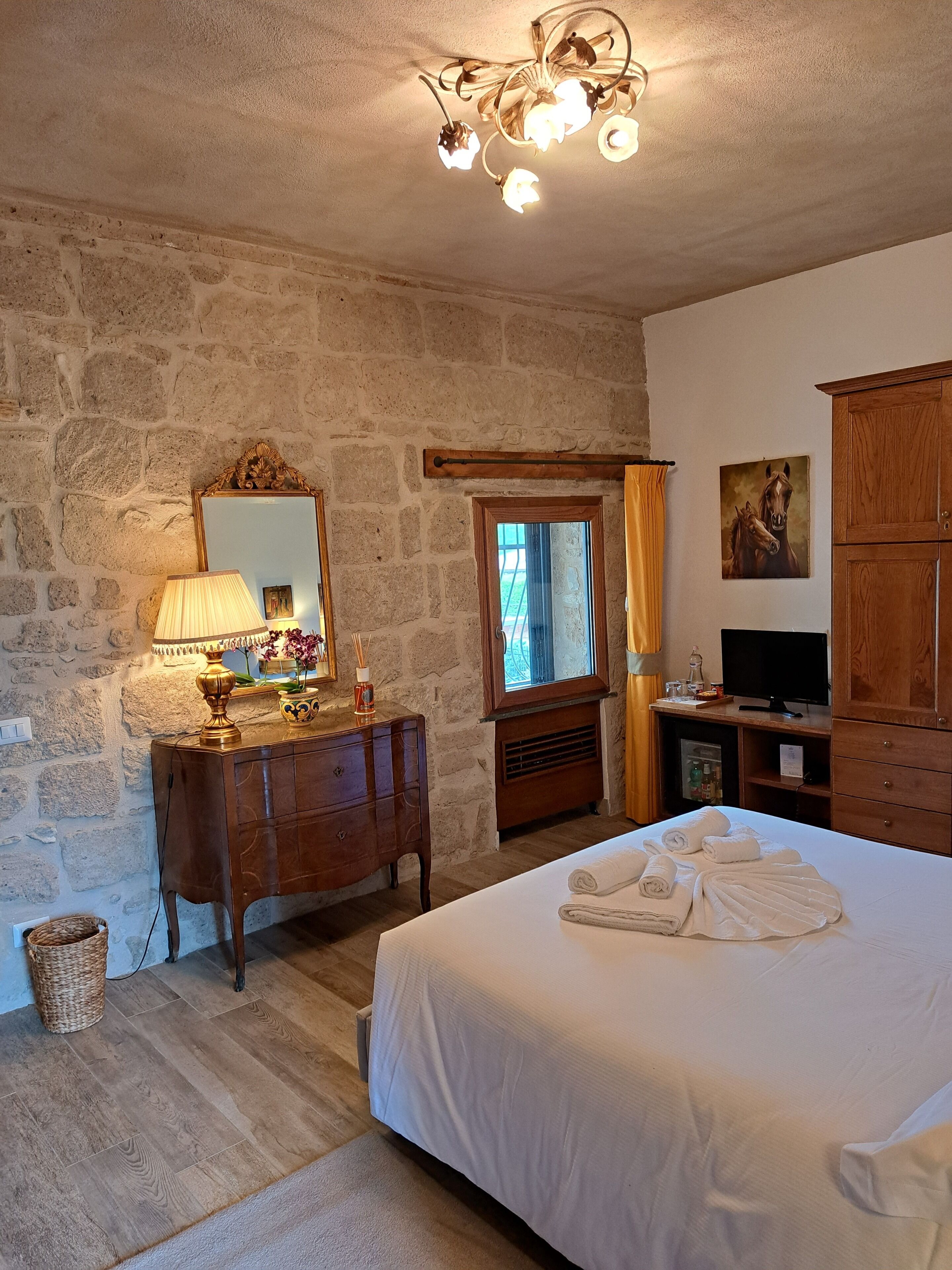 Une chambre avec un mur en pierre, des meubles en bois, un lit avec des serviettes, un miroir, un lustre et un tableau.