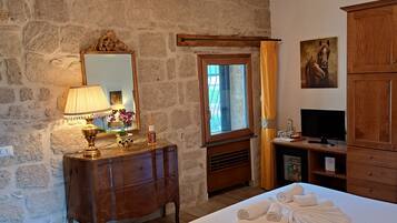 Une chambre avec un mur en pierre, des meubles en bois, un lit avec des serviettes, un miroir, un lustre et un tableau.