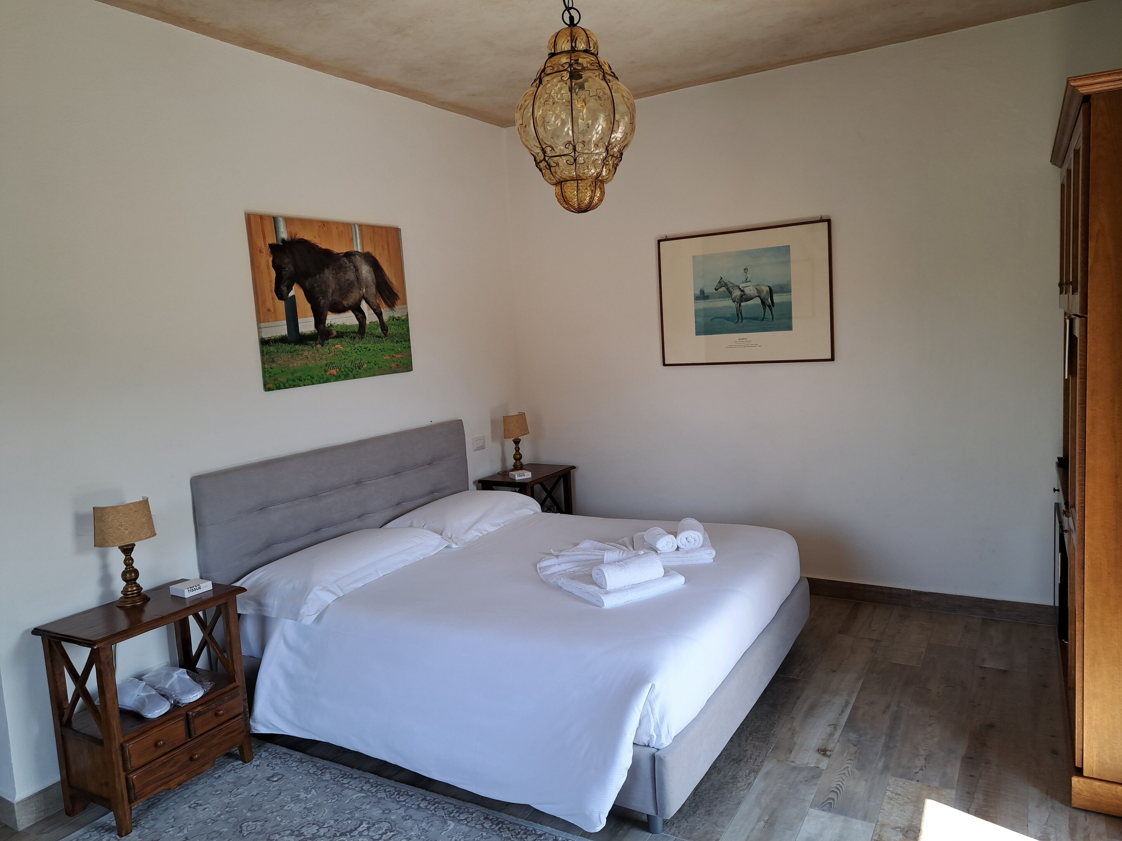 A bedroom with a large bed, two bedside tables, a chandelier, and two framed pictures on the wall.