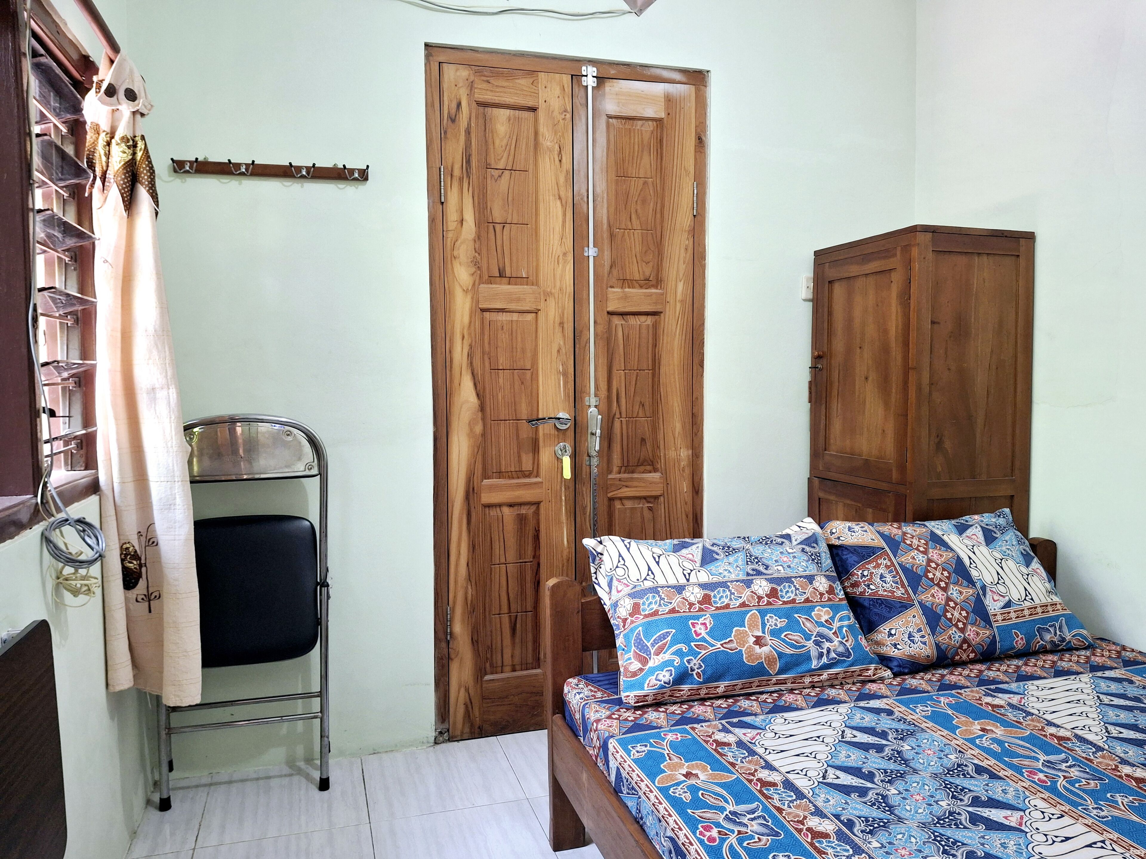 A bedroom with a wooden wardrobe, a metal shelf, a bed with patterned bedding, and a curtain.