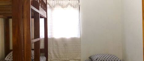 A small, single-room accommodation with bunk beds, a window with a curtain, and an air conditioning unit.