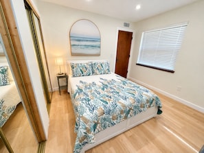 A bedroom with a bed, a nightstand, a mirror, a door, and a window with blinds.