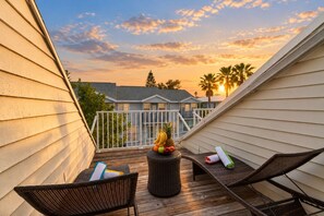 Una terraza de madera con dos sillas de mimbre, una mesita con frutas y una toalla enrollada, y una barandilla que da vista a una zona residencial con casas y palmeras durante el atardecer.
