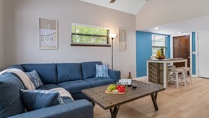 Ein Wohnzimmer mit einem blauen Sofa, einem Holztisch mit Früchten und einer Küche im Hintergrund.