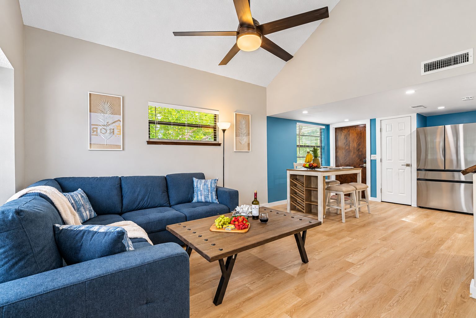 A modern living room with a blue sofa, a wooden coffee table with fruits and a bottle, and a ceiling fan.