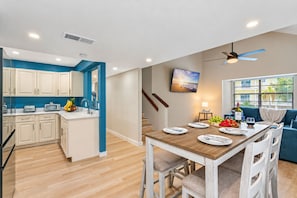 A modern kitchen with a central island, a dining area with a wooden table and chairs, a ceiling fan, and a TV mounted on the wall.