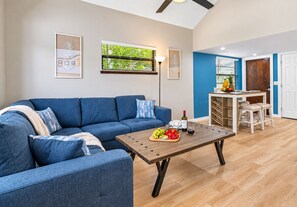 Una sala moderna con un sofá azul, una mesa de centro de madera con frutas y vino, y un comedor con una mesa y sillas de madera.
