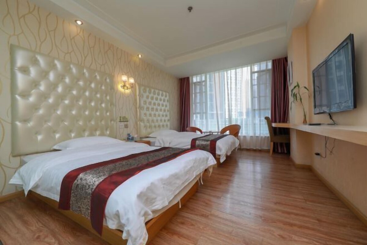 A hotel room with three beds, a TV, a desk, and a large window with curtains.