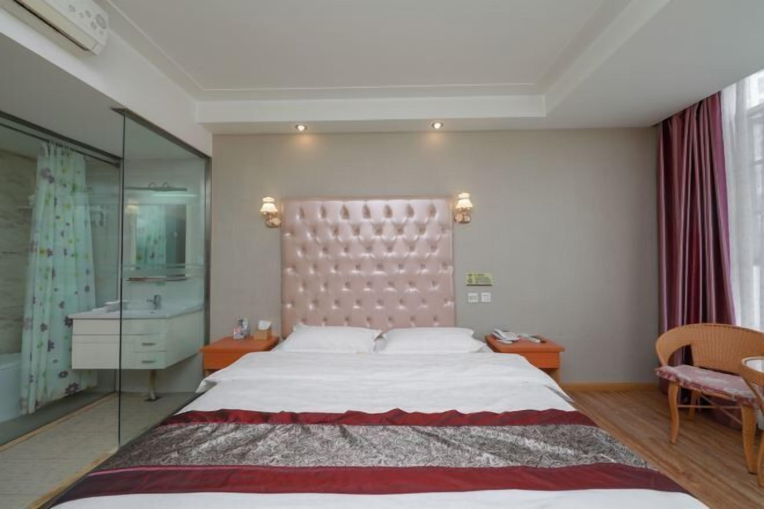Une chambre à coucher avec une tête de lit capitonnée, un lit recouvert d’une couvre-lit à motifs, une table de chevet avec une lampe, un fauteuil et une salle de bain fermée par une paroi en verre.