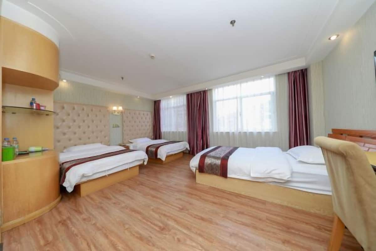 A hotel room with three beds, wooden flooring, a tufted headboard, and a large window with curtains.