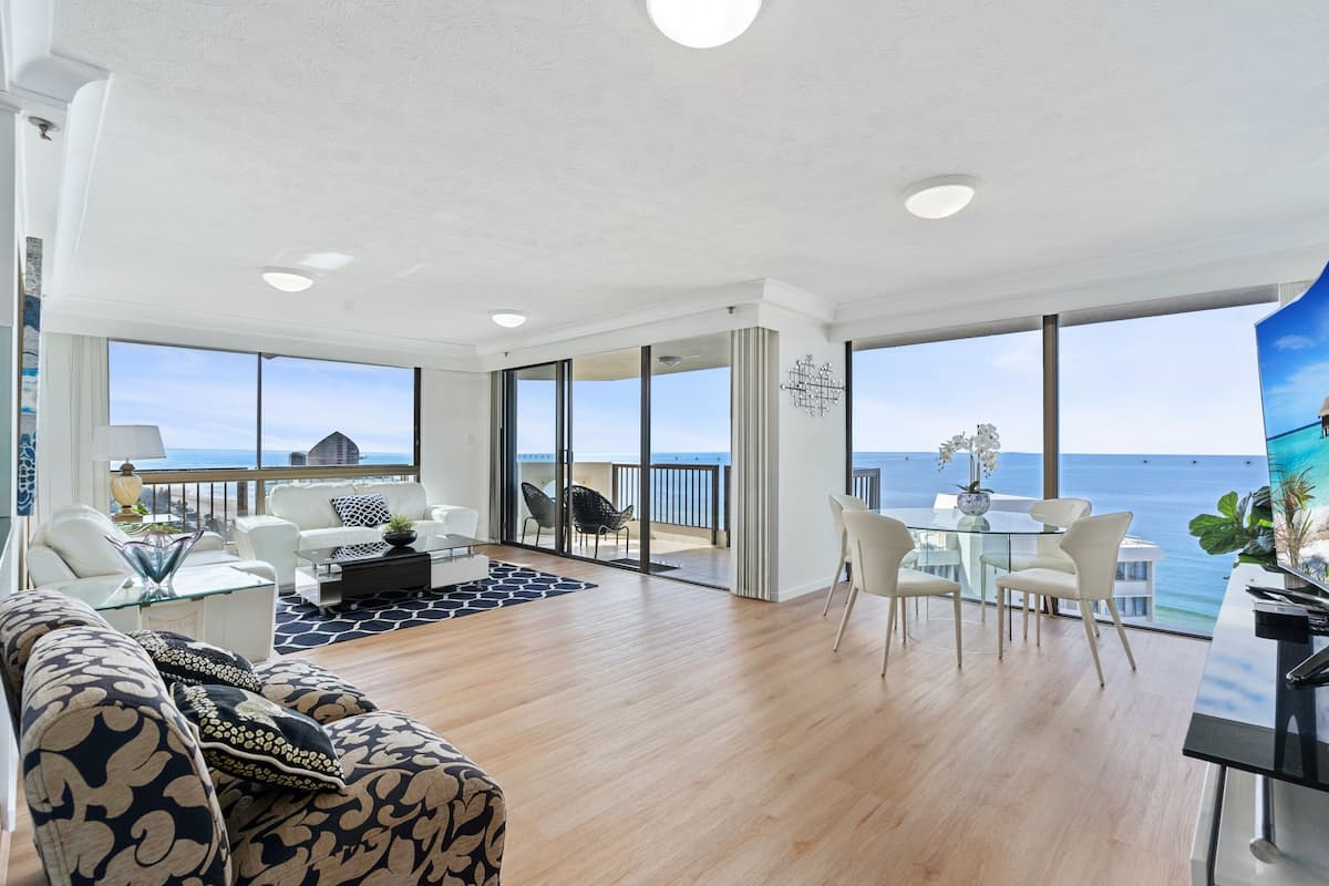 Ampla sala de estar com varanda vista para o mar, mobiliada com sofás brancos, uma mesa de centro de vidro e uma mesa de jantar com cadeiras.