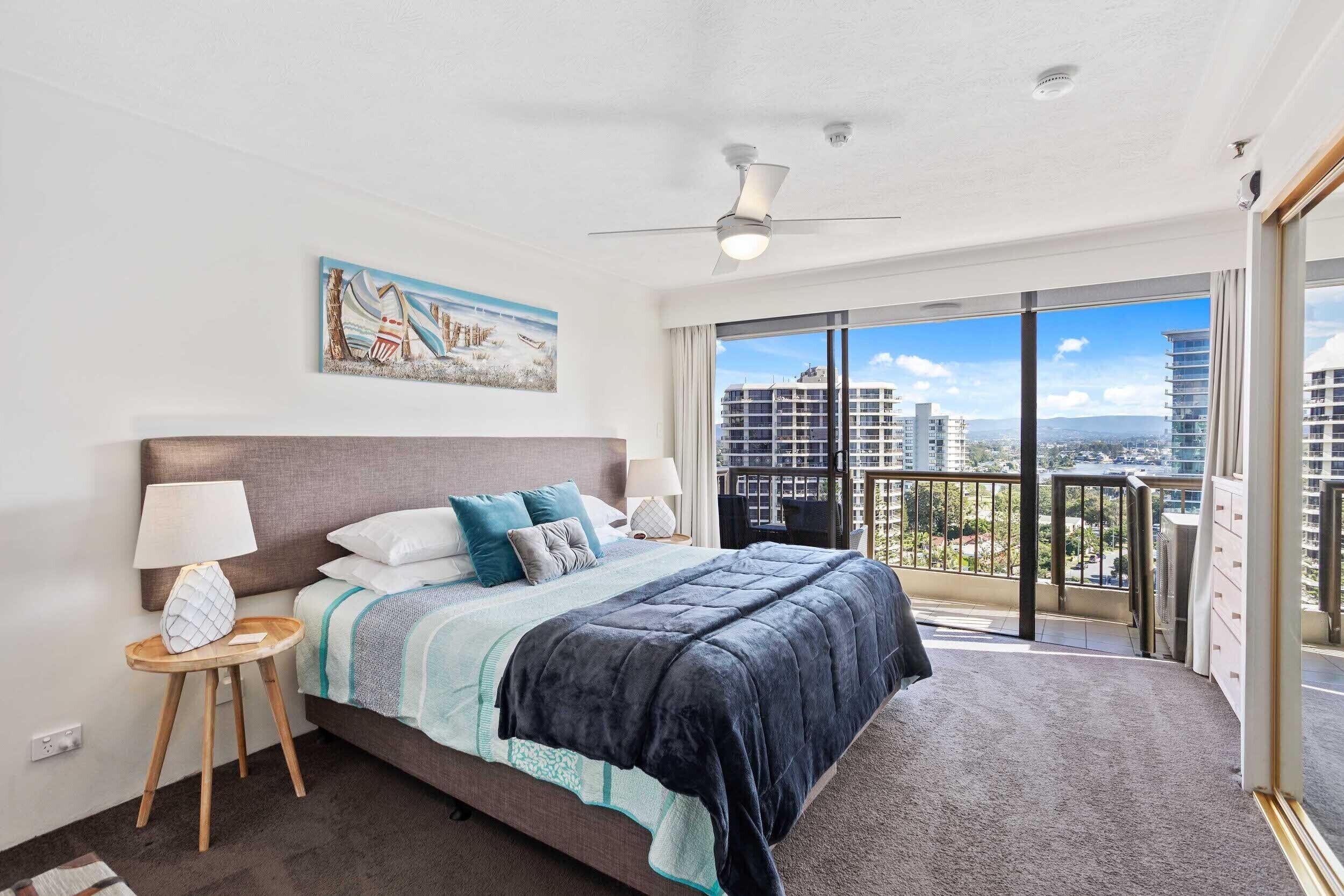 A modern bedroom with a large bed, a ceiling fan, a nightstand with a lamp, and a painting on the wall.