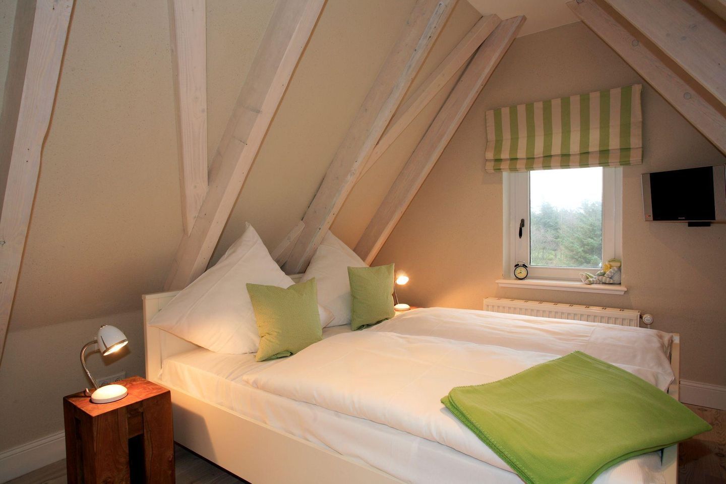 A bedroom with a sloped ceiling, a bed with white bedding, green pillows, a bedside lamp, a window with a striped valance, and a TV mounted on the wall.