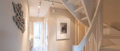 Un hall d’entrée spacieux, doté d’un plancher en bois, d’un escalier blanc et d’un luminaire fixé au mur.