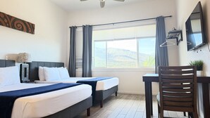 A hotel room with a bed, two bedside tables, a desk, a chair, a ceiling fan, a large window with curtains, and a TV mounted on the wall. - Unit 18 Waena Inn - Maui Private Suite (Wailuku)
