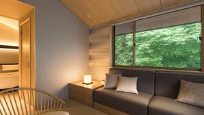 Un dormitorio moderno con techo de madera, un sofá cama con almohadas, una mesita de noche con lámpara y una ventana con vistas a la vegetación.