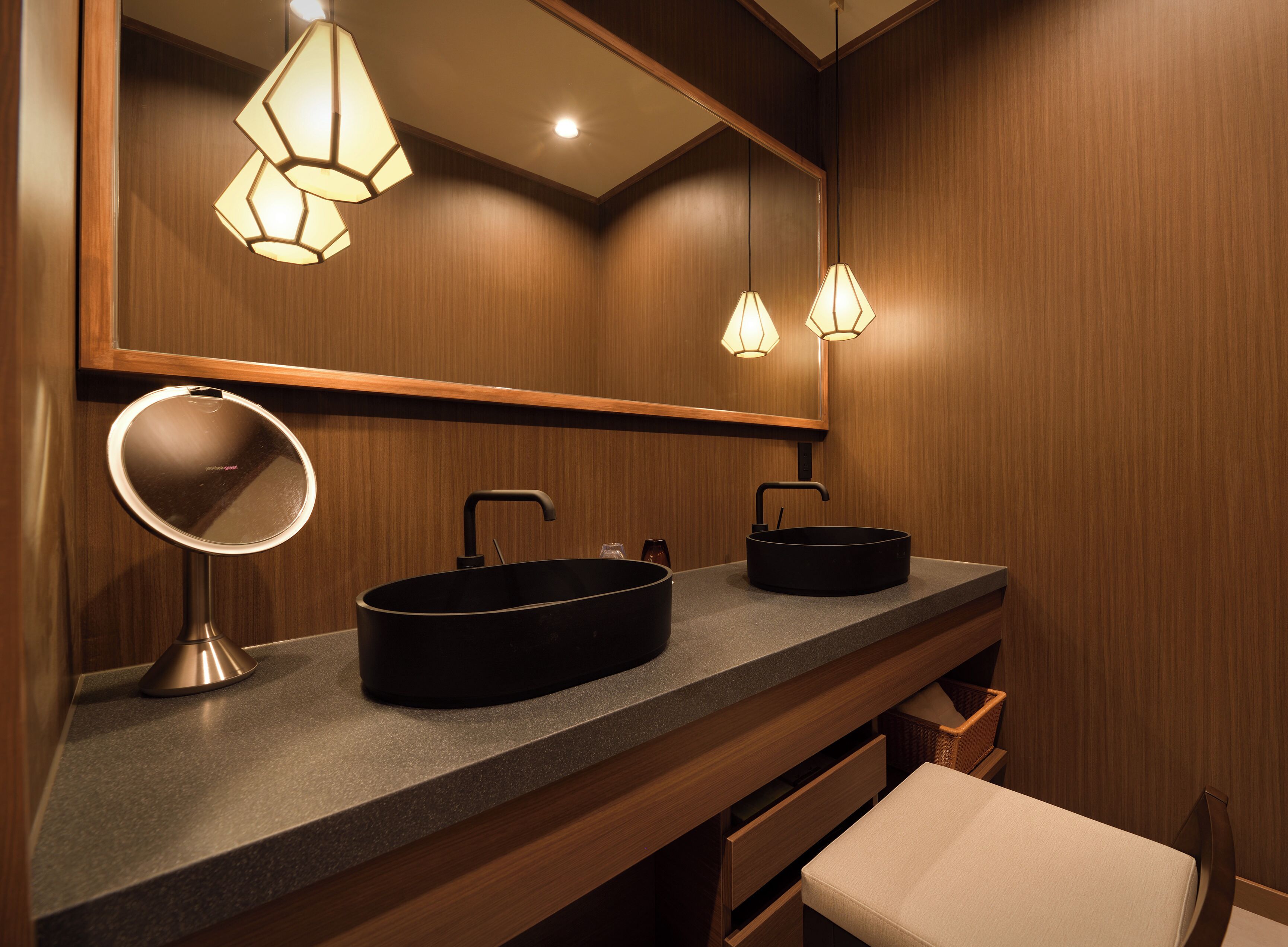A bathroom with a wooden vanity, two black sinks, a round mirror, and pendant lights.