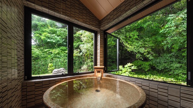 Un baño moderno con una bañera de hidromasaje grande y circular, ventanas amplias con vistas a vegetación exuberante y un taburete de madera en el centro.