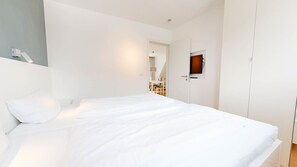 A bedroom with a large bed, white walls, a door with a small window, and a light fixture above the bed.