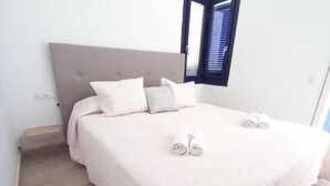 A neatly made bed with a grey headboard, white bedding, and a small window with blinds.