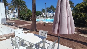 An outdoor seating area with white tables and chairs, a pink umbrella, and a pool in the background.