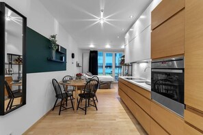 A modern kitchen with a built-in oven, a dining area with a table and chairs, and a view of the outside through large windows.