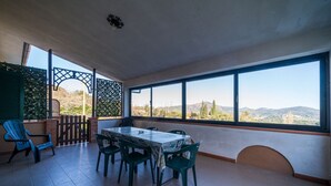 Un pati espaiós amb taula i cadires, grans finestres que ofereixen vistes a les colines, i una zona coberta amb una pantalla de gelosia.