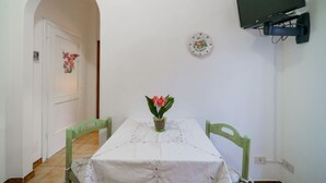 A dining area with a table set for two, a single flower vase, and a television mounted on the wall.