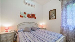 A bedroom with a bed, two bedside tables, a nightstand, a wall-mounted air conditioner, and a window with floral curtains.