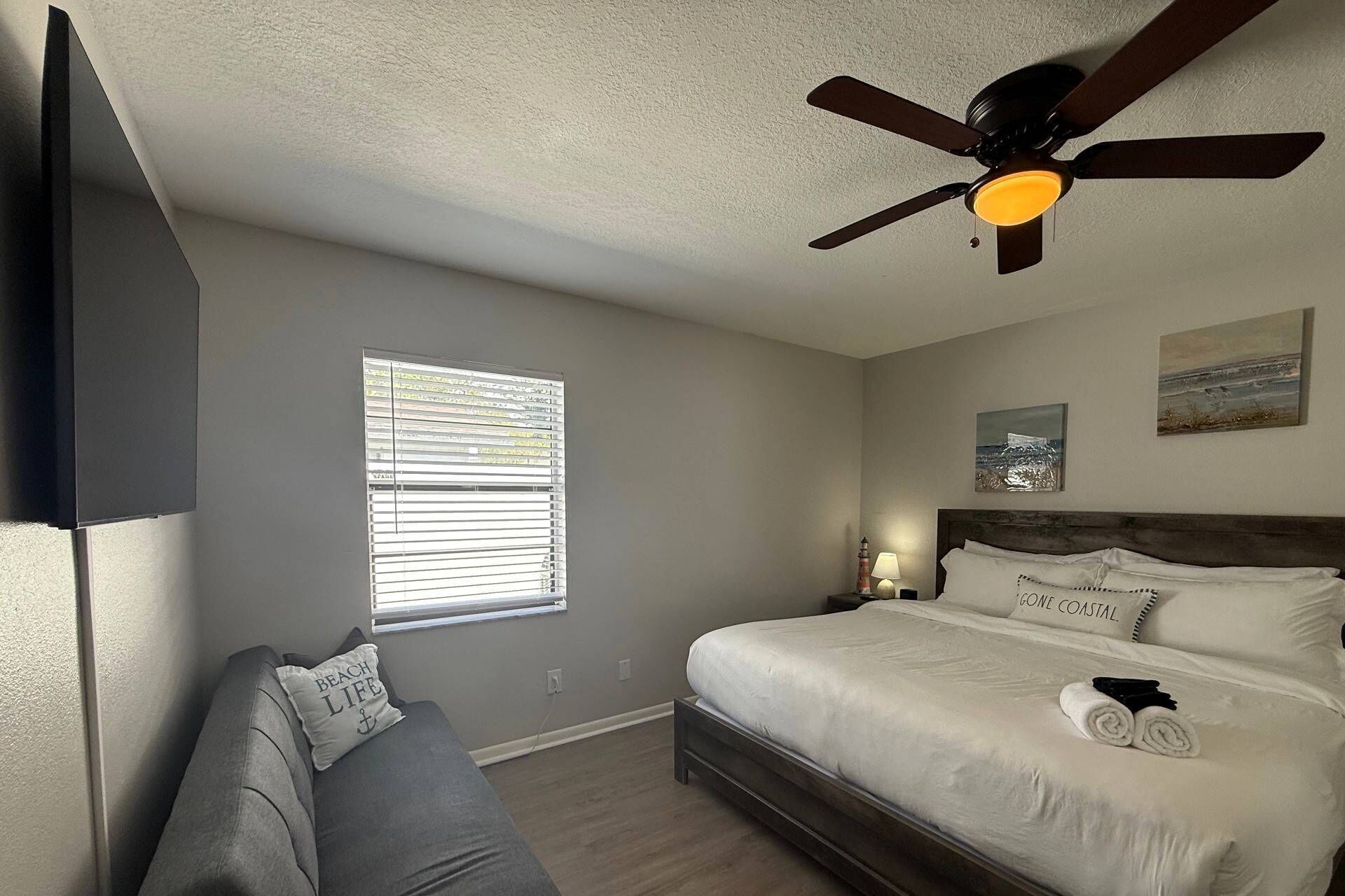A bedroom with a bed, a ceiling fan, a window with blinds, a bedside table with a lamp, and a painting on the wall.