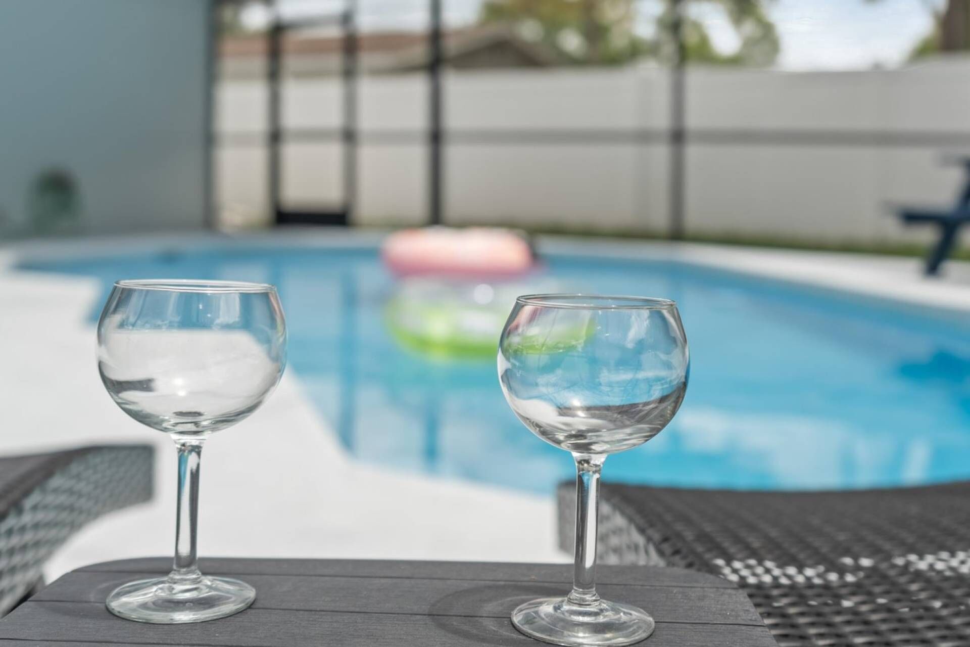 Two empty wine glasses on a wooden table by a swimming pool.