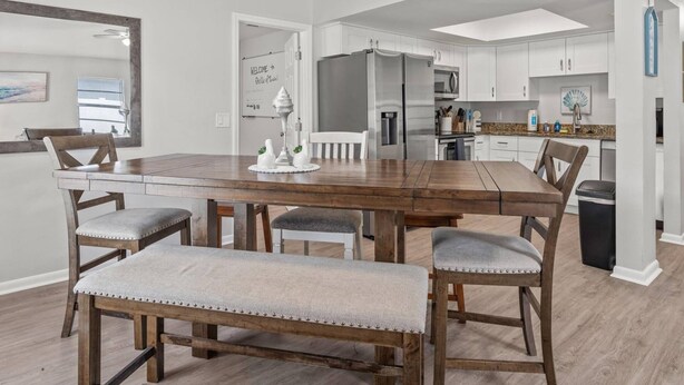 Un comedor con una mesa de madera y cuatro sillas, un banco, un refrigerador y una cocina al fondo.