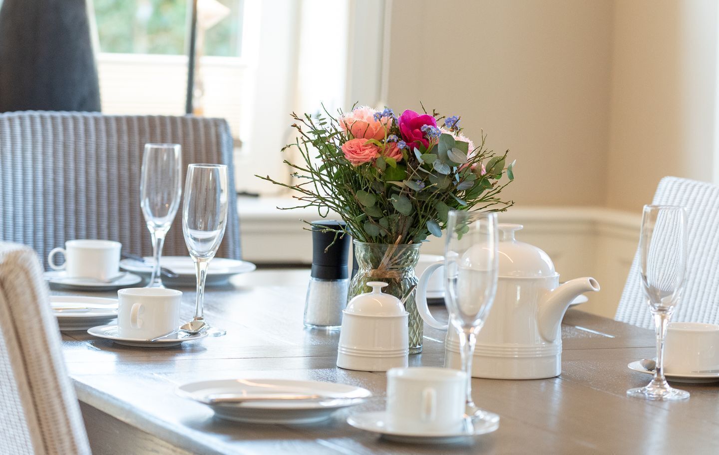 Una mesa de comedor dispuesta con tazas y platillos blancos, una tetera y un florero con flores.