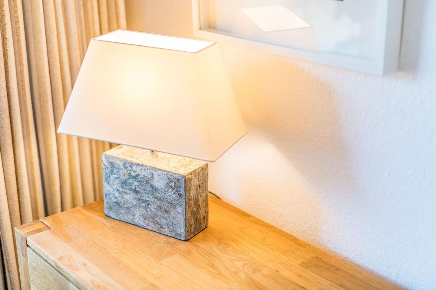 Une lampe de table avec un abat-jour blanc, posée sur une table en bois, devant un mur orné de stores verticaux.