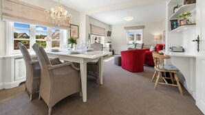 Un comedor con una mesa blanca dispuesta para cuatro personas, rodeada de sillas de mimbre. Se observa un sofá rojo y una silla de madera al fondo.