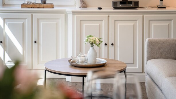 A round table with a white vase and glassware, a grey sofa, and white cabinetry in the background.