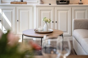 Une table ronde avec un vase blanc et des verres, un canapé gris et des meubles blancs en arrière-plan.