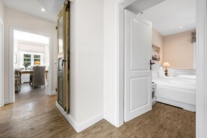 Un couloir mène à une chambre à coucher qui contient un lit, une table de chevet et une lampe fixée au mur. Il y a une porte sur la gauche et une fenêtre au fond.