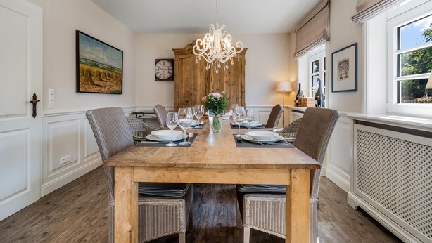 Een eetkamer met een houten tafel gedekt voor zes personen, omringd door rieten stoelen, een kroonluchter en een klok aan de muur.