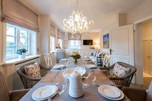 A dining room with a long table set for six, surrounded by wicker chairs, a chandelier, and a kitchen in the background. - Haus Borelör Whg. 1 EG - EG im Haus Börelör, 3-zimmerwohnung auf ca. 80 m² im EG für bis zu 4 (Sylt)