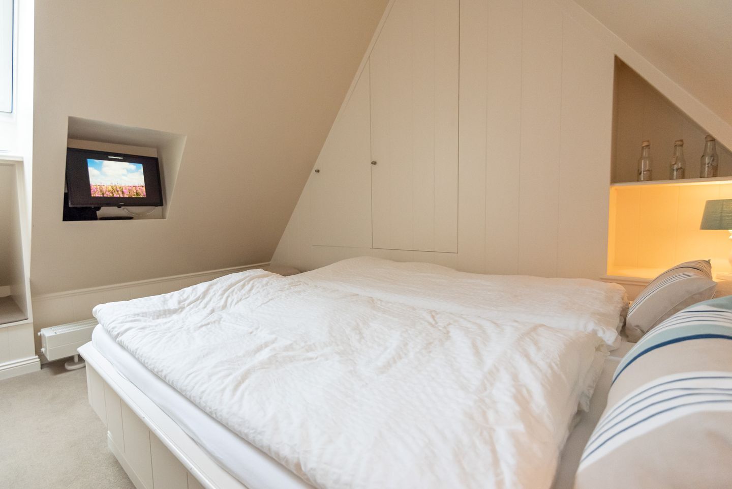 A bedroom with a bed, a television mounted on the wall, and a shelf with bottles.