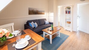 Un salón acogedor con un sofá gris oscuro, una mesa de centro de madera y una mesa de comedor dispuesta con platos y vasos. Se ve una estantería al fondo.