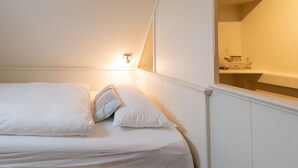 A bedroom with a bed, pillows, a nightstand, and a mirror reflecting a sink and a towel.